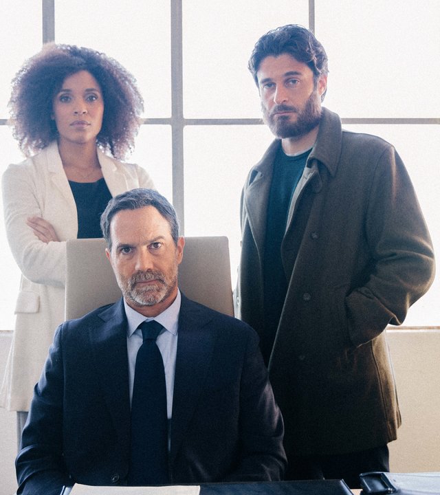 "Survivors - Zurück ins Leben": Sylvie (Stéfi Celma) und ihr Mann Luca (Lino Guanciale) stehen hinter dessen bestem Freund Stefano (Fausto Sciarappa), der auf einem Stuhl hinter seinem Schreibtisch in einem Büro der Werft sitzt. Sie schauen angestrengt nach vorne.