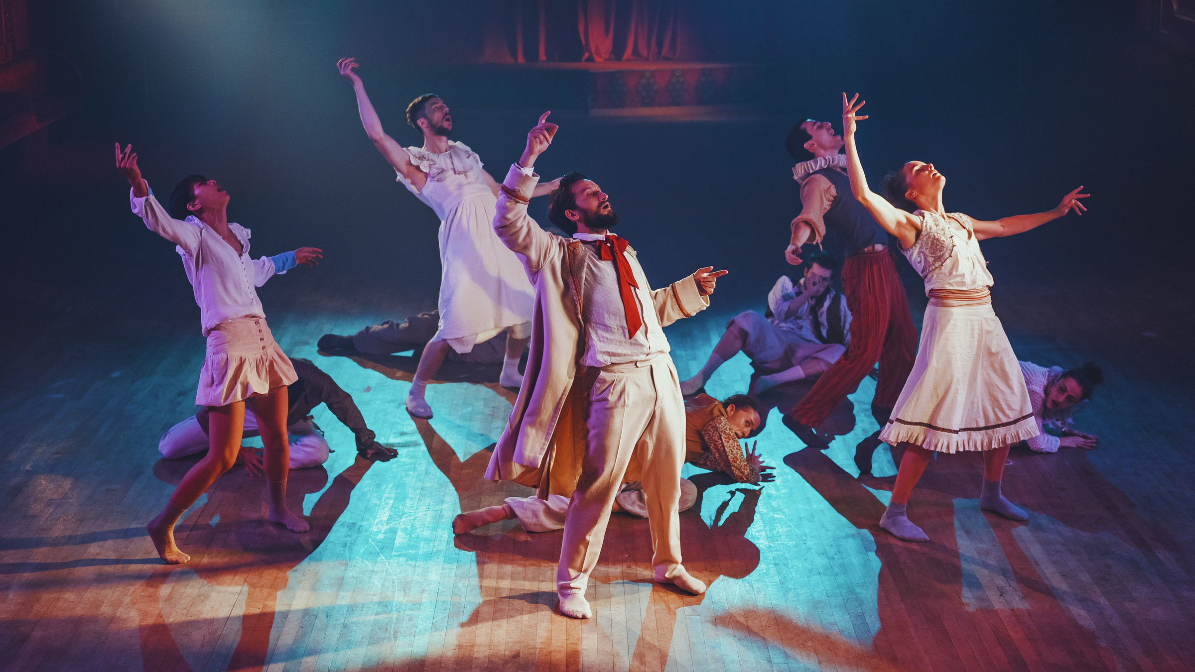 "Clowns": Mehrere Personen befinden sich auf einer Bühne mit hellbraunem Holzboden: Fünf stehen breitbeinig in einer leicht zurückgelehnten Haltung mit erhobenem rechten Arm, sechs liegen räkelnd am Boden. Alle tragen unterschiedliche Kostüme.