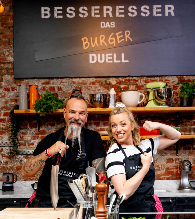 "besseresser: Das Burger-Duell": Ralf und Zora stehen vor einer Klinkerwand mit Küchendeko. Ralf hält ein großes Messer in der Hand, das er auf einem Schneidebrett abstützt, Zora ahmt eine Bodybuilder-Pose nach. Über ihnen hängt ein Schild mit dem Sendungstitel.