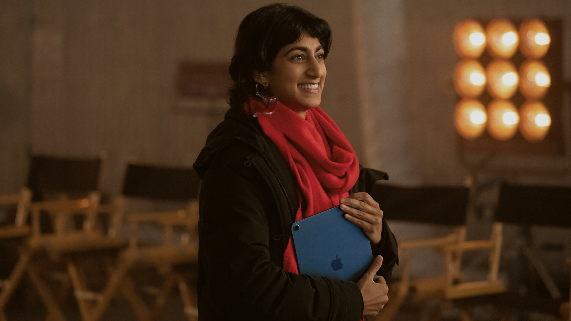 "Bupkis - Alles geht schief": Monica (Sunita Mani) steht am Filmset. Im Hintergrund stehen aufgereiht viele Stühle und die Scheinwerfer für den Dreh leuchten. Monika hält ein Notebook in der Hand, ihr roter Schal strahlt und sie schaut lächelnd zur Seite.