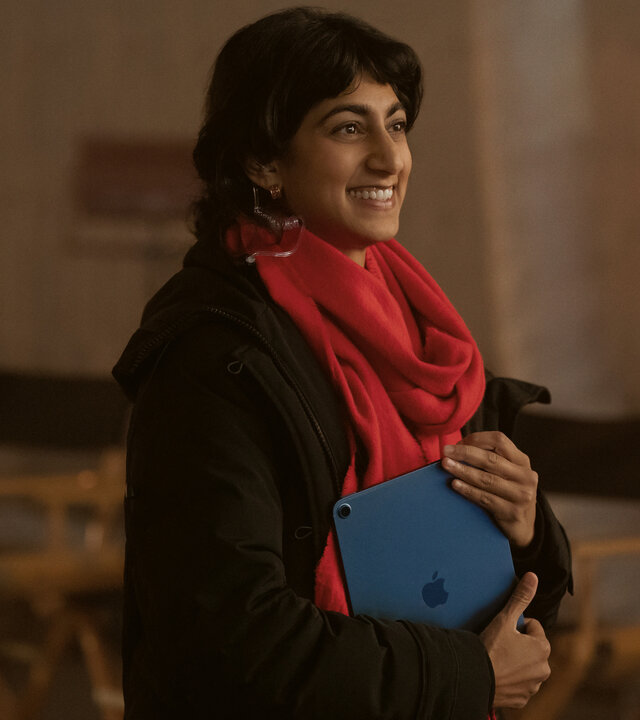 "Bupkis - Alles geht schief": Monica (Sunita Mani) steht am Filmset. Im Hintergrund stehen aufgereiht viele Stühle und die Scheinwerfer für den Dreh leuchten. Monika hält ein Notebook in der Hand, ihr roter Schal strahlt und sie schaut lächelnd zur Seite.