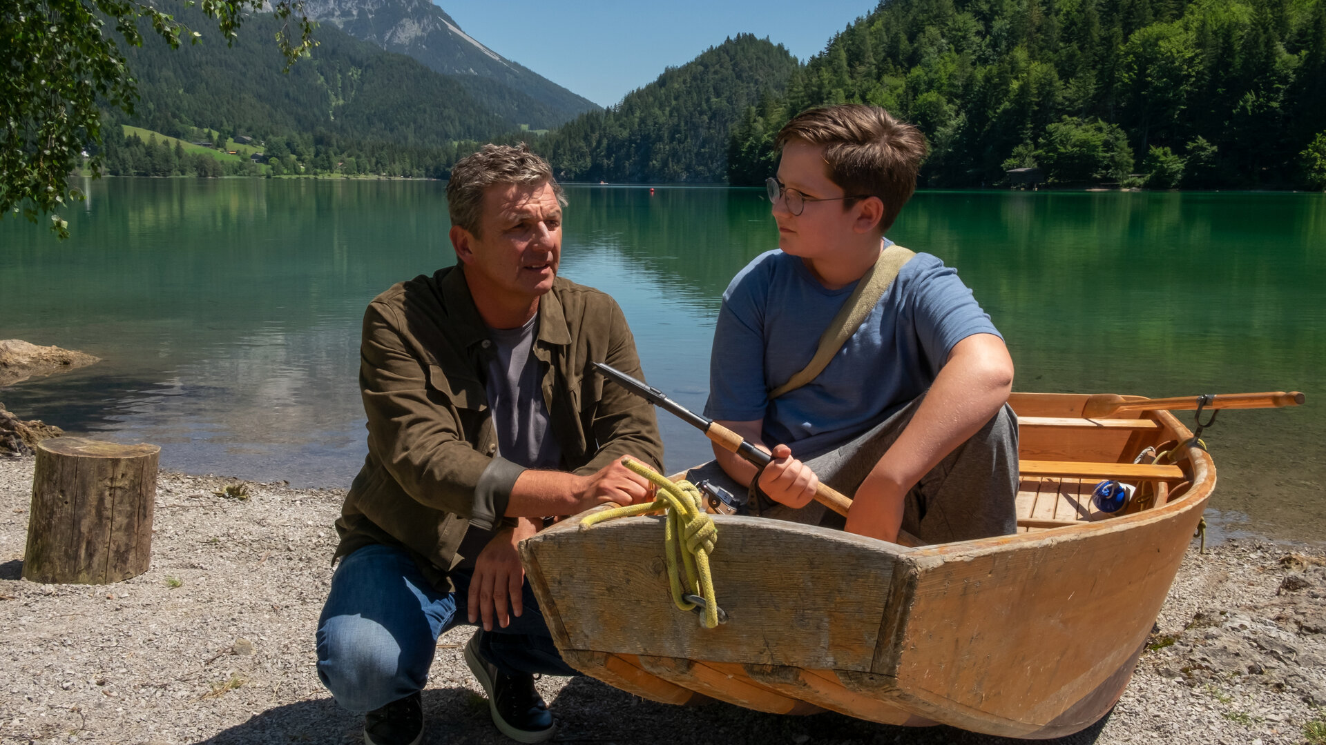 "Der Bergdoktor: Eine schwere Last": Am Rand des Ufers eines Bergsees sitzt Sammy Federer (Emil Knapp) in einem Ruderboot und hält eine Angel in der Hand. Neben dem Boot kniet Dr. Martin Gruber (Hans Sigl) und blickt nachdenklich.