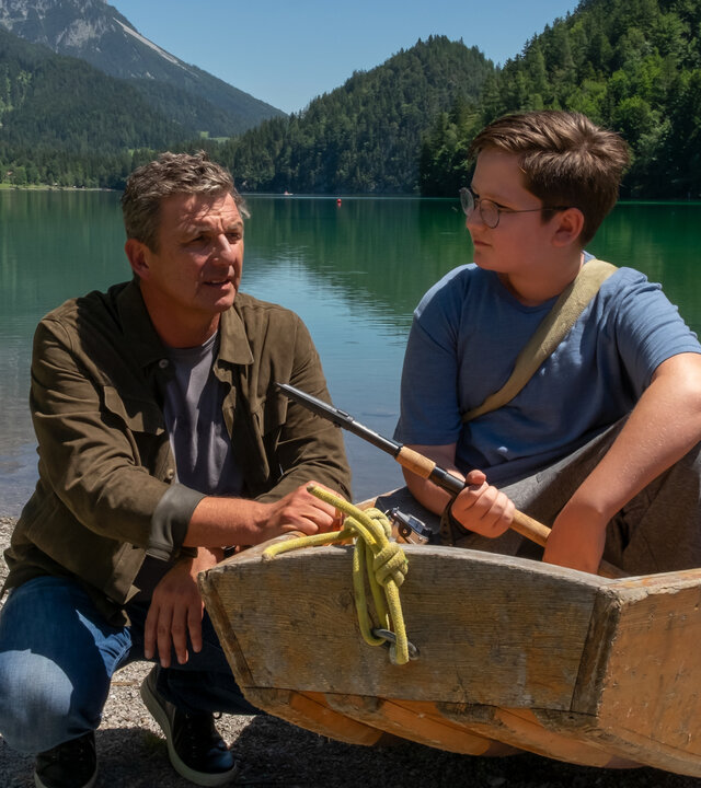 "Der Bergdoktor: Eine schwere Last": Am Rand des Ufers eines Bergsees sitzt Sammy Federer (Emil Knapp) in einem Ruderboot und hält eine Angel in der Hand. Neben dem Boot kniet Dr. Martin Gruber (Hans Sigl) und blickt nachdenklich.