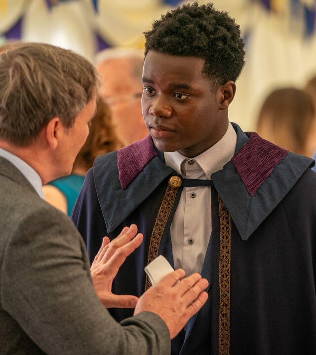 "Boarders - Welcome to St. Gilbert's - Lunch & Loyalty": Bernard Ashford (Derek Riddell) und Jaheim (Josh Tedeku) stehen sich in einem mit Wimpelketten geschmückten Großraumzelt gegenüber. Jaheim ist festlich mit einem Umhang gekleidet. Bernard, der nur von hinten zu sehen ist, spricht mit ihm und gestikuliert. Der Schulleiter hält ein Stück Papier in der Hand. Hinter ihnen befinden sich weitere Personen (Komparsen).