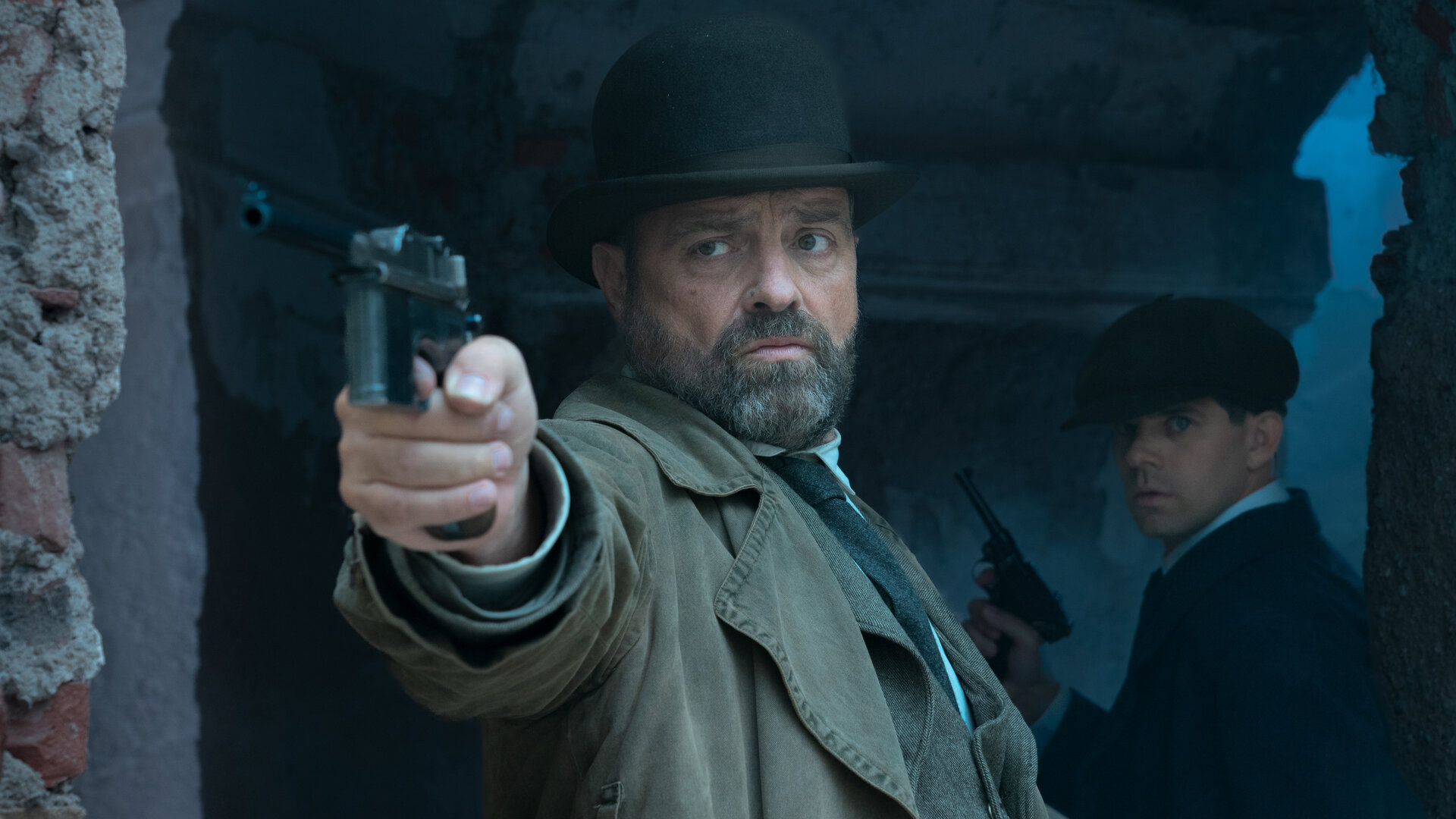 "Vienna Blood - Mephisto Teil 2": Oskar Rheinhardt (Juergen Maurer) steht im düsteren Durchgang eines ruinenartigen Gebäudes und richtet seine Pistole auf einen Punkt hinter der Kamera, hinter ihm steht Haussmann (Josef Ellers), ebenfalls mit einer Pistole in der Hand.