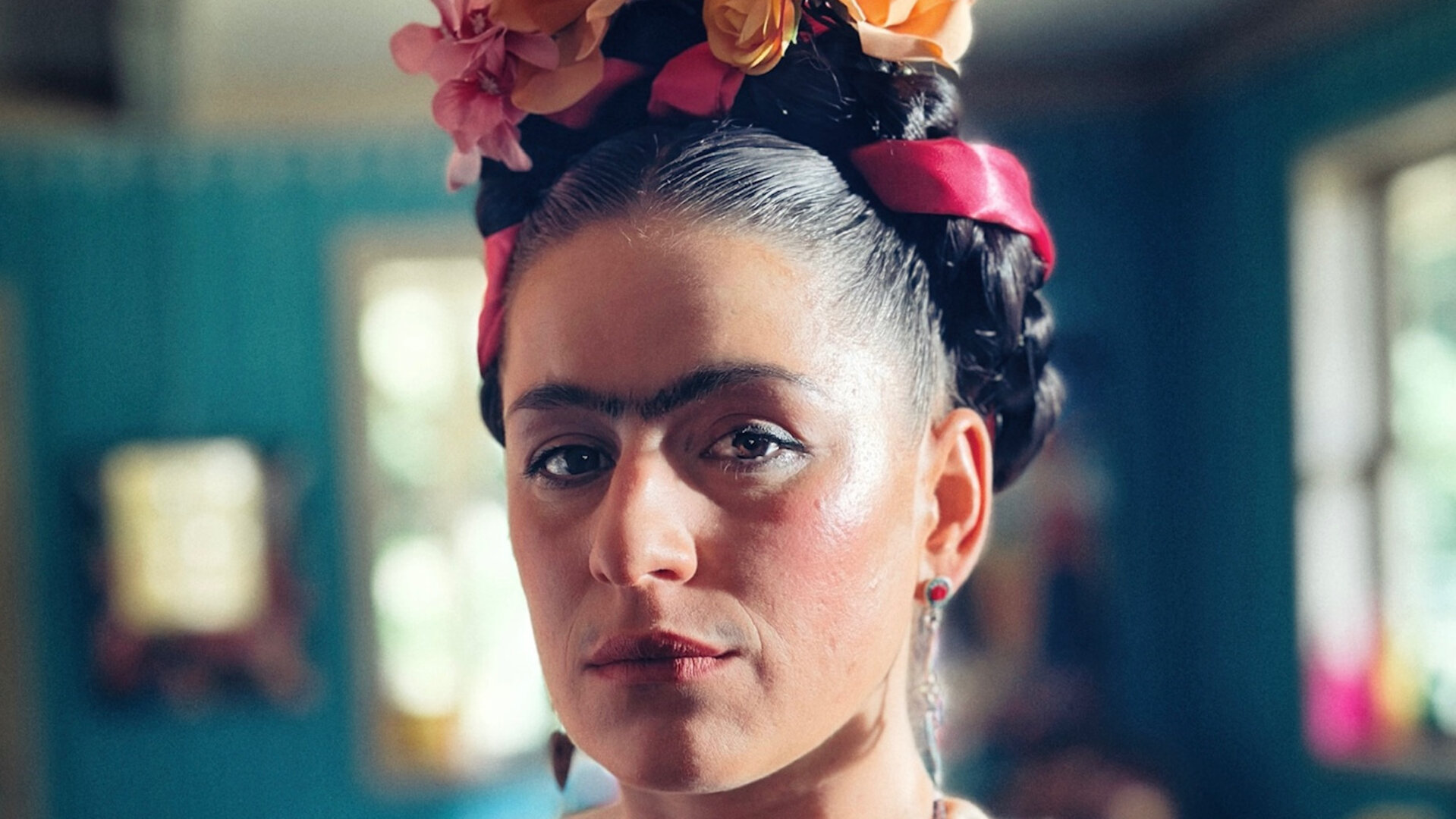 "Sketch History - Gute Fridas, schlechte Fridas": Frida Kahlo (Lou Strenger) steht in einem sehr bunten Raum. Sie ist stark geschminkt und ihre Augenbrauen gehen ineinander über. Sie trägt eine bunte mexikanische Tracht mit einem üppigen Blumenkranz in den Haaren.