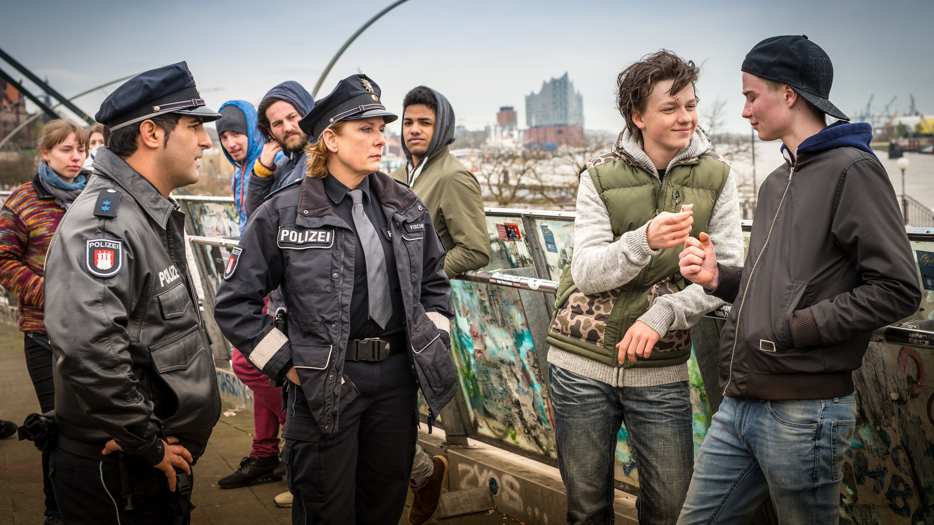"Notruf Hafenkante - Die Nervensäge": Mit fassungslosem Gesichtsausdruck stehen die Polizisten Tarik Coban (Serhat Cokgezen) und Claudia Fischer (Janette Rauch) vor Dustin Neumann (Jeremias Meyer). Der lehnt lässig an einem mit Graffiti besprühten Geländer neben einem Jugendlichen (Komparse) und hält grinsend einen Geldschein in der rechten Hand.