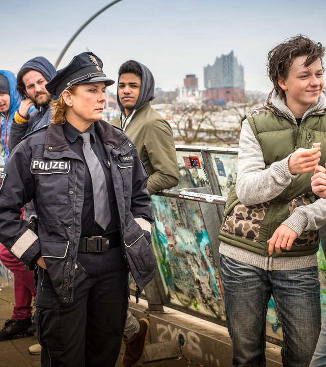 "Notruf Hafenkante - Die Nervensäge": Mit fassungslosem Gesichtsausdruck stehen die Polizisten Tarik Coban (Serhat Cokgezen) und Claudia Fischer (Janette Rauch) vor Dustin Neumann (Jeremias Meyer). Der lehnt lässig an einem mit Graffiti besprühten Geländer neben einem Jugendlichen (Komparse) und hält grinsend einen Geldschein in der rechten Hand.