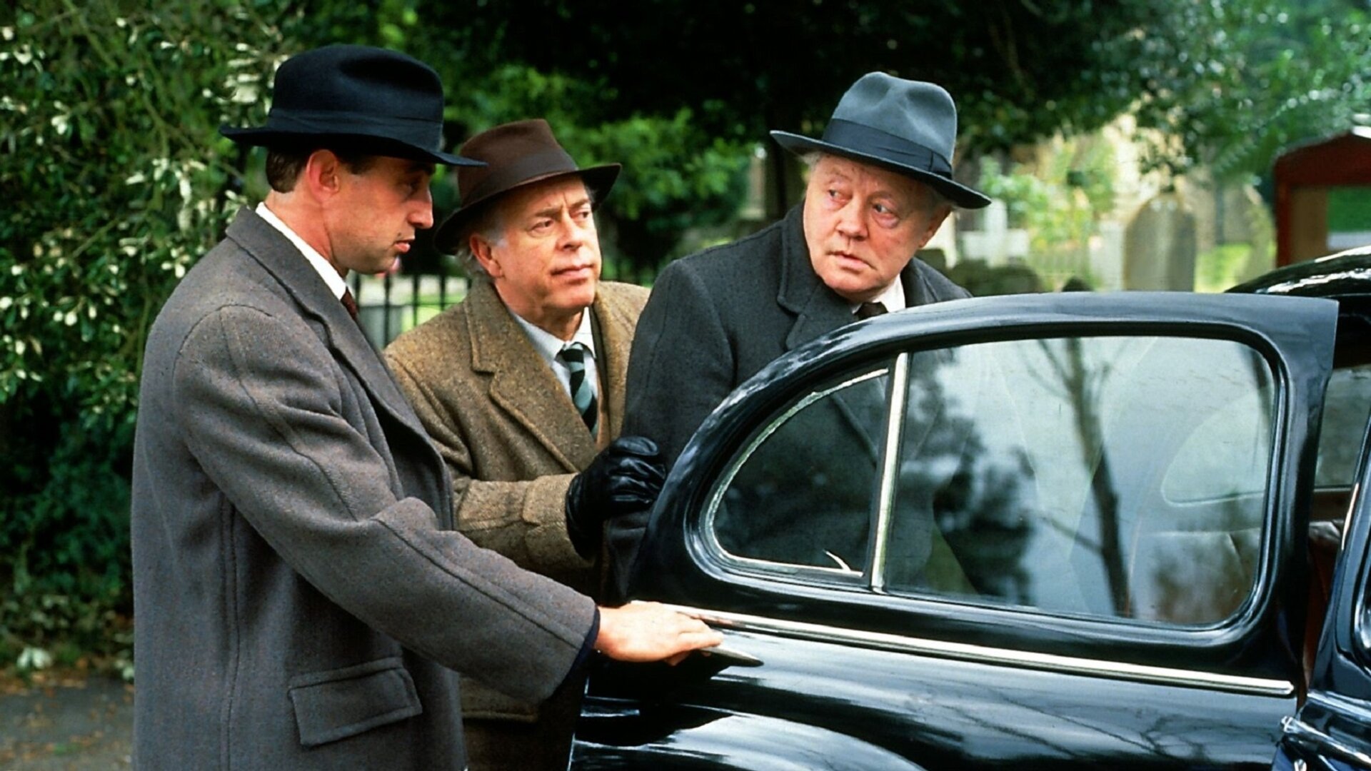 "Fünf Freunde und ein Gipsbein (2)": Ein Mann (Unbekannt), Pottersham (Clive Swift) und Terry-Kane (Dudley Sutton) tragen alle Hüte und Mäntel und stehen dabei neben einem schwarzen Auto. Die Tür des Autos ist geöffnet. Pottersham (Clive Swift) hat seine Hand an dem Arm von Terry-Kane (Dudley Sutton), der den dritten Mann, der seine Hand an der geöffneten Autotür hat, skeptisch anschaut.