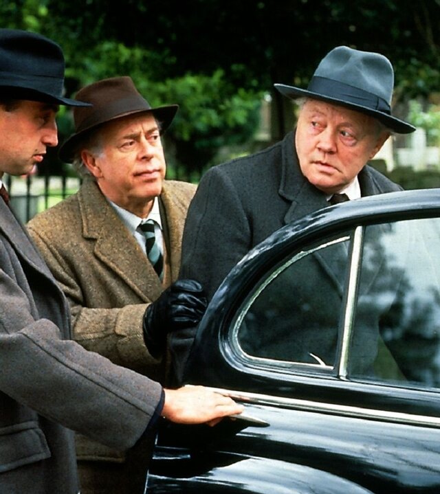 "Fünf Freunde und ein Gipsbein (2)": Ein Mann (Unbekannt), Pottersham (Clive Swift) und Terry-Kane (Dudley Sutton) tragen alle Hüte und Mäntel und stehen dabei neben einem schwarzen Auto. Die Tür des Autos ist geöffnet. Pottersham (Clive Swift) hat seine Hand an dem Arm von Terry-Kane (Dudley Sutton), der den dritten Mann, der seine Hand an der geöffneten Autotür hat, skeptisch anschaut.