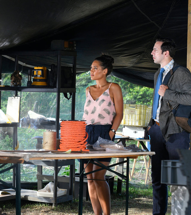 "Death in Paradise - Tod des Professors": DS Cassell (Joséphine Jobert) und DI Parker (Ralf Little) stehen Ed Lancer (Luke Pasqualino) in einem Zelt voller Ausrüstung gegenüber und schauen ihn konzentriert an.