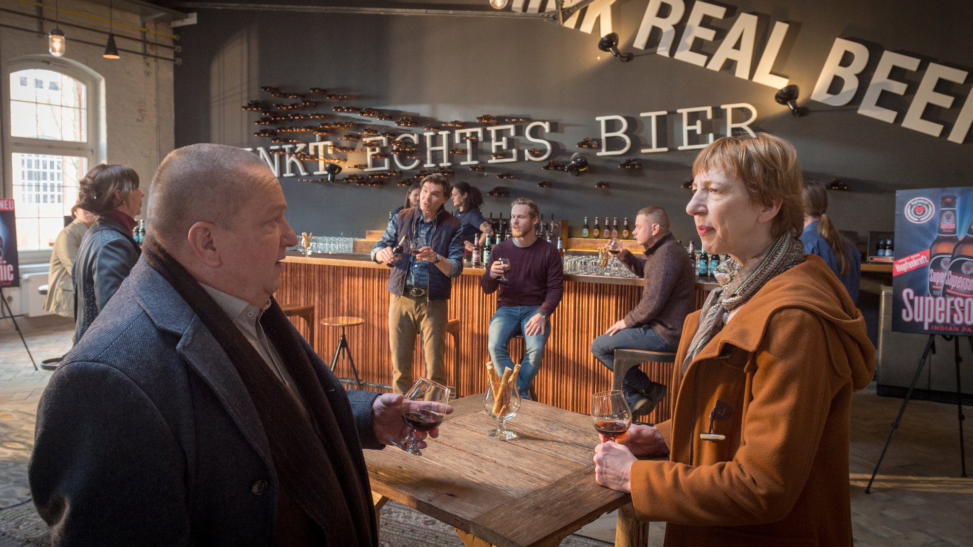 "SOKO Wismar – Der Bierflüsterer": Jan Reuter (Udo Kroschwald) und Helene Sturbeck (Katharina Blaschke) stehen an einem Tisch und halten ein Glas Bier in der Hand. Im Hintergrund befindet sich ein langer Tresen, an dem mehrere Besucher und Mitarbeiter der Kneipe zu sehen sind (Komparsen).