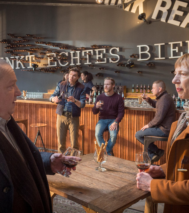 "SOKO Wismar – Der Bierflüsterer": Jan Reuter (Udo Kroschwald) und Helene Sturbeck (Katharina Blaschke) stehen an einem Tisch und halten ein Glas Bier in der Hand. Im Hintergrund befindet sich ein langer Tresen, an dem mehrere Besucher und Mitarbeiter der Kneipe zu sehen sind (Komparsen).