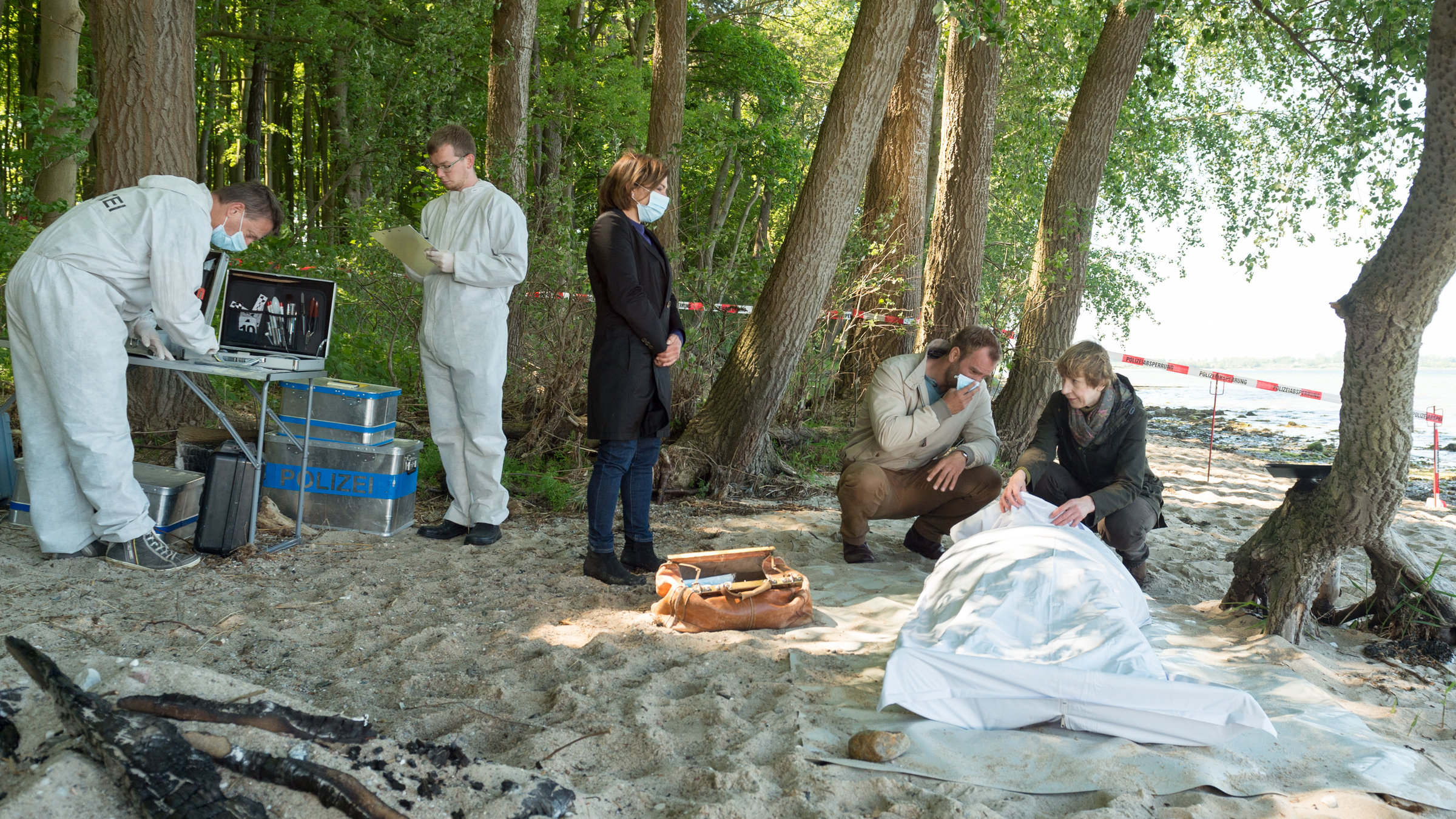 "SOKO Wismar – Todesstrafe": An einer Strandbucht zwischen Bäumen ist ein Tatort abgesperrt. Helene Sturbeck (Katharina Blaschke) kniet auf dem Boden hebt leicht das Leichentuch an. Neben ihr kniet Lars Pöhlmann (Dominic Boeer), der sich einen Mundschutz vor das Gesicht hält. Etwas abseits steht Katrin Börensen (Claudia Schmutzler), ebenfalls mit Mundschutz. Außerdem sind die Kollegen von der KTU (Komparsen) bereits bei der Arbeit.