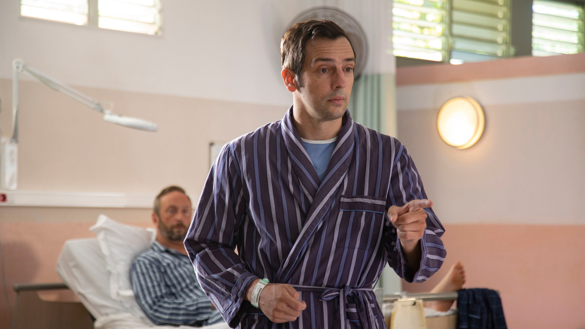 "Death in Paradise - Tödliche Nachtschicht": DI Parker (Ralf Little) steht in einem Morgenmantel vor einem Krankenbett und deutet auf etwas in der Ferne.
