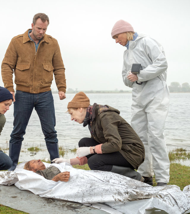 "SOKO Wismar – Die Sprache der Wunden": Melanie Reusdorf (Anne Baus) liegt in einem Leichensack auf einer Moorwiese. Um sie herum hocken Karoline Joost (Nike Fuhrmann) und Helene Sturbeck (Katharina Blaschke). Ebenfalls bei ihr stehen Lars Pöhlmann (Dominic Boeer) und Roswitha Prinzler (Silke Matthias). Im Hintergrund flutet Wasser die Moorlandschaft.