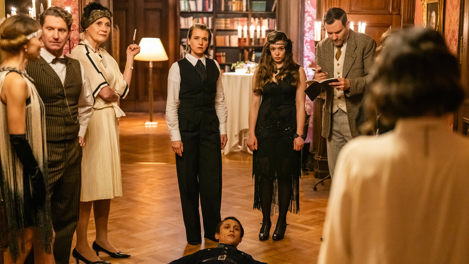 "SOKO Köln - 1926": Vanessa Haas (Kerstin Landsmann), Amira Mahdi (Aylin Ravanyar) und Jonas Fischer (Lukas Piloty) stehen nebeneinander in einem Saal. Vanessa und Amira blicken schockiert zu Lilly Funke (Tatjana Kästel), die mit dem Rücken zur Kamera auf der anderen Seite des Raumes steht. Zwischen ihr und ihren Kollegen liegt Jora Temur (Madieu Ulbrich) auf dem Parkett und sieht ernst zu Lilly. An der Seite stehen nebeneinander Brigitte Keilmann (Alexandra von Schwerin), Max Balberg (Max Rothbart) und Katja Temur (Inga Lessmann). Brigitte Keilmann hat die Arme vor dem Körper verschränkt und sieht ebenfalls ernst zu Lilly. Max Balberg und Katja Temur unterhalten sich.