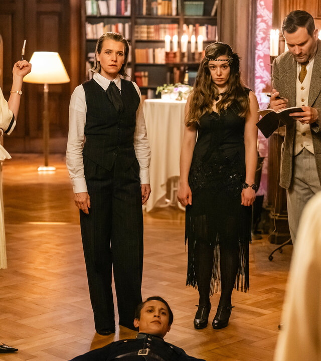 "SOKO Köln - 1926": Vanessa Haas (Kerstin Landsmann), Amira Mahdi (Aylin Ravanyar) und Jonas Fischer (Lukas Piloty) stehen nebeneinander in einem Saal. Vanessa und Amira blicken schockiert zu Lilly Funke (Tatjana Kästel), die mit dem Rücken zur Kamera auf der anderen Seite des Raumes steht. Zwischen ihr und ihren Kollegen liegt Jora Temur (Madieu Ulbrich) auf dem Parkett und sieht ernst zu Lilly. An der Seite stehen nebeneinander Brigitte Keilmann (Alexandra von Schwerin), Max Balberg (Max Rothbart) und Katja Temur (Inga Lessmann). Brigitte Keilmann hat die Arme vor dem Körper verschränkt und sieht ebenfalls ernst zu Lilly. Max Balberg und Katja Temur unterhalten sich.