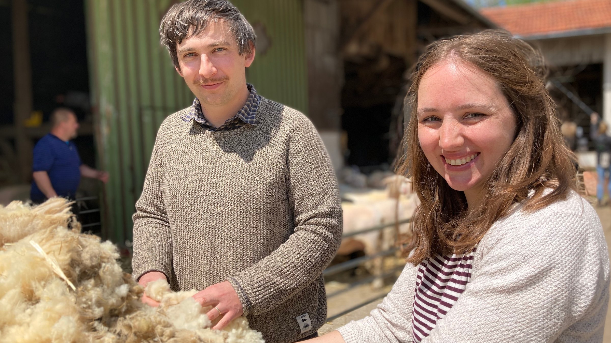 "plan b: fair und flauschig": Ruth Werwai und Marten Wellbrock stehen vor einem Lagerschuppen und greifen in einen Wollehaufen.