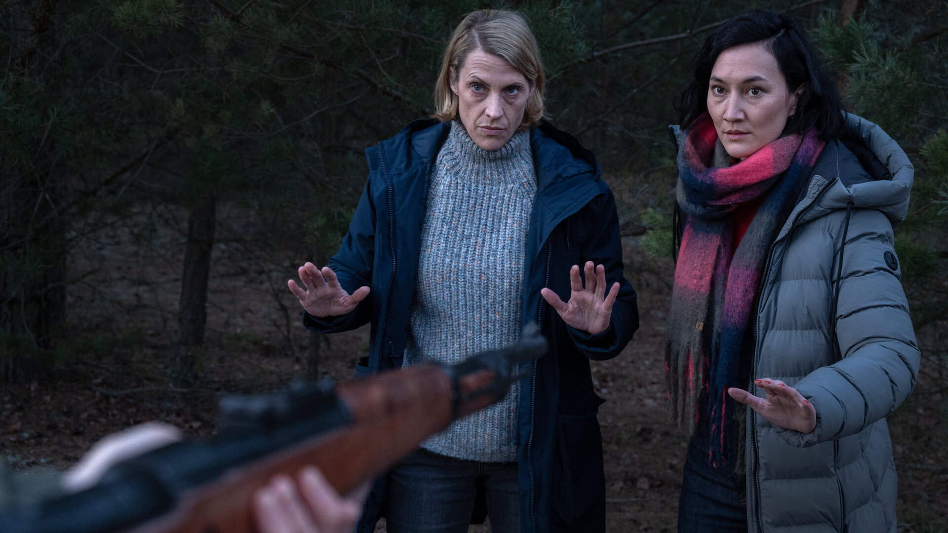 "SOKO Potsdam - Nachts im Wald": Im dichten Wald stehen Tamara Meurer  (Anja Pahl) und Pauline Hobrecht (Agnes Decker), die ihre Hände abwehrend hochhalten. Tamara Meurer blickt ernst und konzentriert nach vorn, neben ihr steht Pauline Hobrecht. Vor beiden ist ein Gewehrlauf auf sie gerichtet.