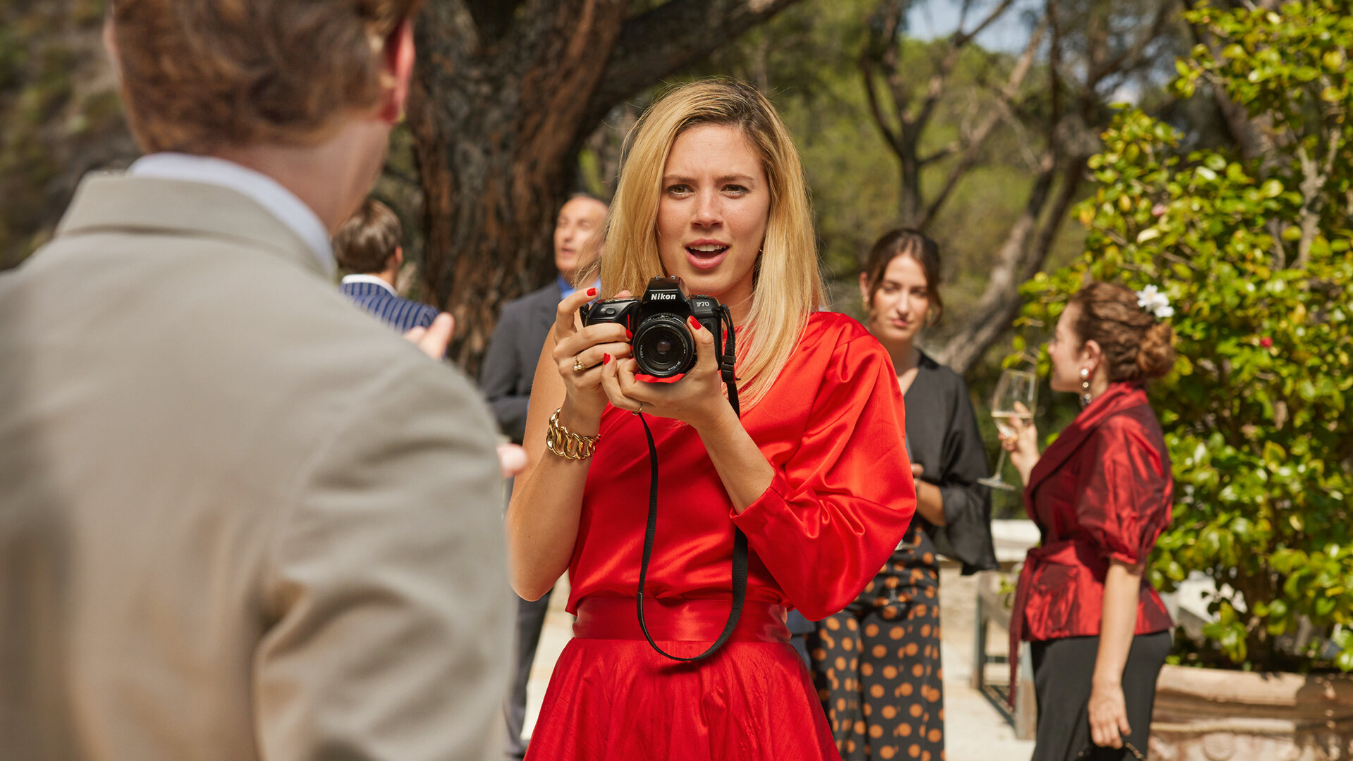 "Maxima - Die Begegnung": Auf einer Terrasse: Maxima (Delfina Chaves) lässt die Kamera sinken und sucht Blickkontakt zu dem Prinzen Willem-Alexaner (Martijn Lakemeier). Im Hintergrund befinden sich weitere Personen (Komparsen).