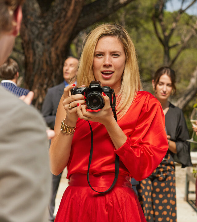 "Maxima - Die Begegnung": Auf einer Terrasse: Maxima (Delfina Chaves) lässt die Kamera sinken und sucht Blickkontakt zu dem Prinzen Willem-Alexaner (Martijn Lakemeier). Im Hintergrund befinden sich weitere Personen (Komparsen).
