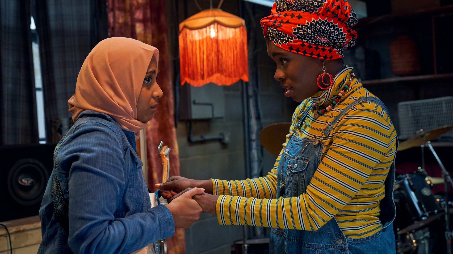 "We are Lady Parts - Erdenbürgerinnen": Amina (Anjana Vasan) und Bisma (Faith Omole) stehen sich in einem dunklen Raum mit Vorhängen gegenüber und halten sich an den Händen. Amina trägt ein rosafarbenes Kopftuch, eine blaue Jacke und hat eine E-Gitarre umgehängt. Bisma trägt ein gelb gestreiftes Oberteil und ein gemustertes Tuch um den Kopf. Im Hintergrund steht ein Schlagzeug.