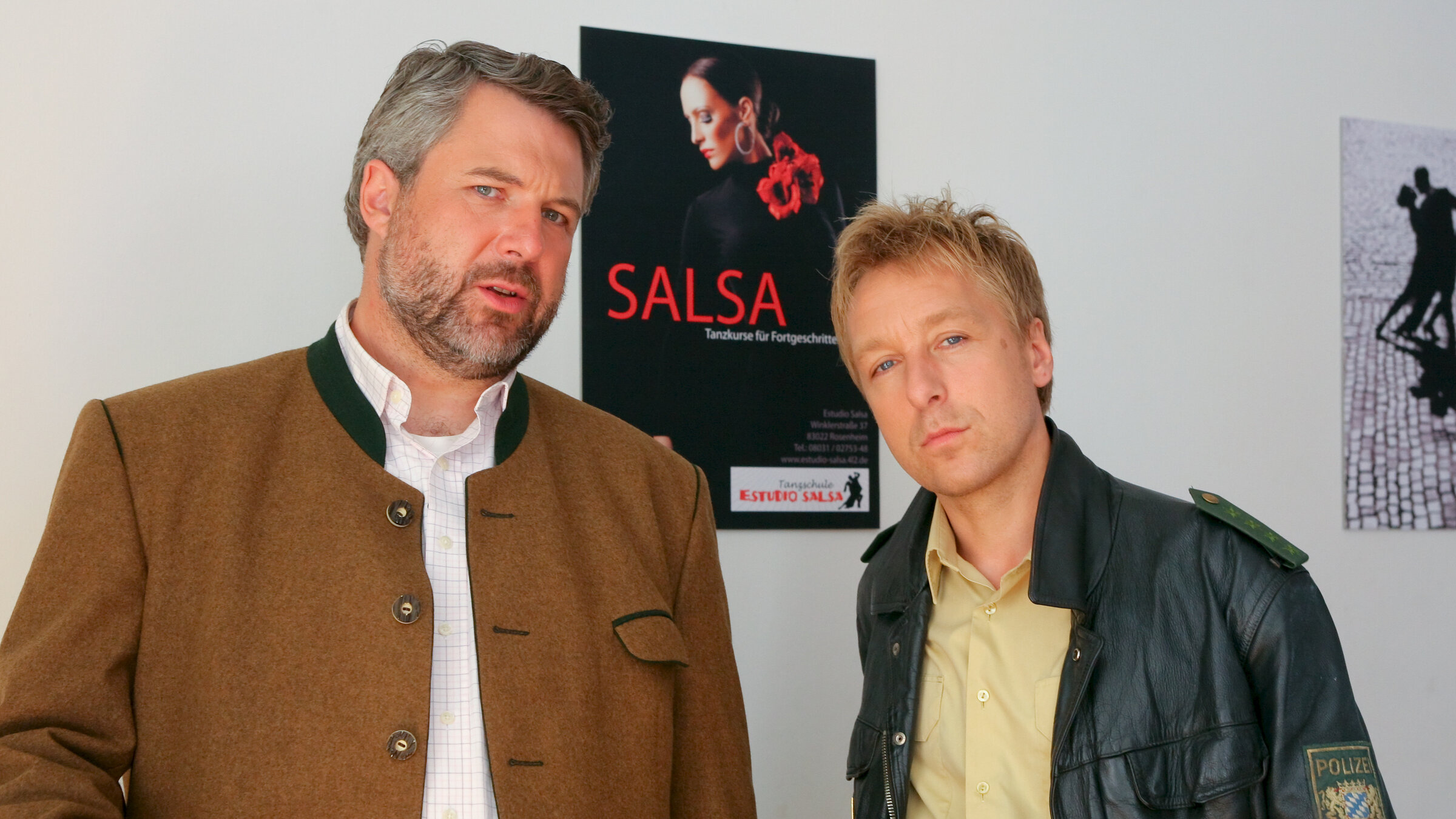 "Die Rosenheim-Cops - Ausgetanzt": Die Rosenheimer Ermittler Stadler (Dieter Fischer) und Mohr (Max Müller) beobachten etwas. Sie stehen vor einer weißen Wand mit Postern. Auf dem Poster hinter ihnen ist eine Frau in schwarzer Kleidung und roten Blumen abgebildet, darunter steht in rot "SALSA".