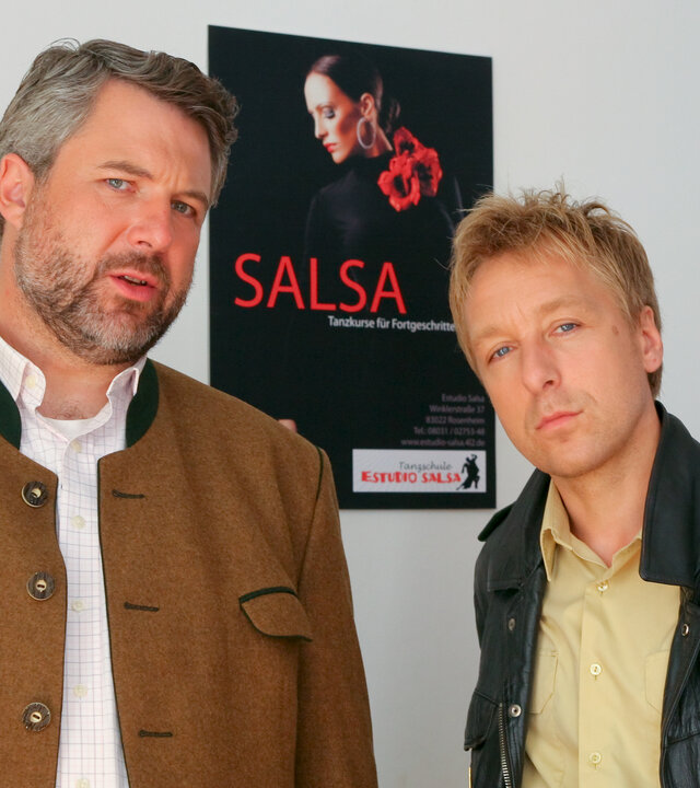 "Die Rosenheim-Cops - Ausgetanzt": Die Rosenheimer Ermittler Stadler (Dieter Fischer) und Mohr (Max Müller) beobachten etwas. Sie stehen vor einer weißen Wand mit Postern. Auf dem Poster hinter ihnen ist eine Frau in schwarzer Kleidung und roten Blumen abgebildet, darunter steht in rot "SALSA".