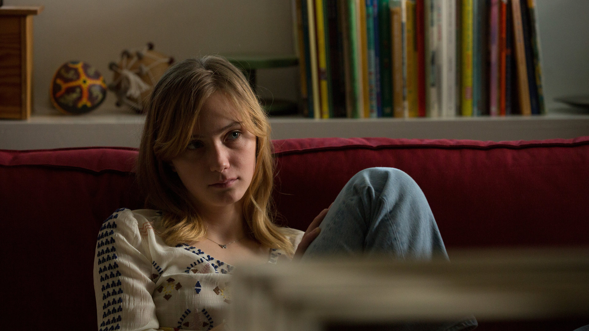 "SAFE - Episode 8": Nellie (Carla Hüttermann) sitzt auf einem Sofa und sieht in die Ferne. Im Hintergrund sind Bücher und diverse Dekorationsgegenstände zu sehen.