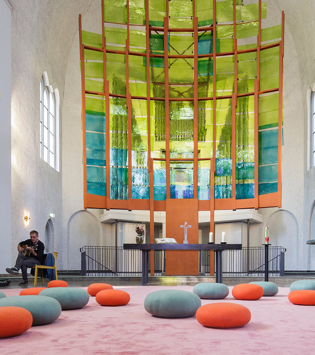 "Evangelischer Gottesdienst: Es ist, was es ist":  Auf dem Boden des lichtdurchfluteten Innenraums der Genezarethkirche in Berlin-Neukölln liegen graue und orangefarbene Sitzkissen. In der hinteren Bildmitte ist ein großes, in Türkis und Gelb gestaltetes Kirchenfenster zu sehen. Im Altarraum sitzt ein Mann mit übergeschlagenem Bein auf einem Stuhl.