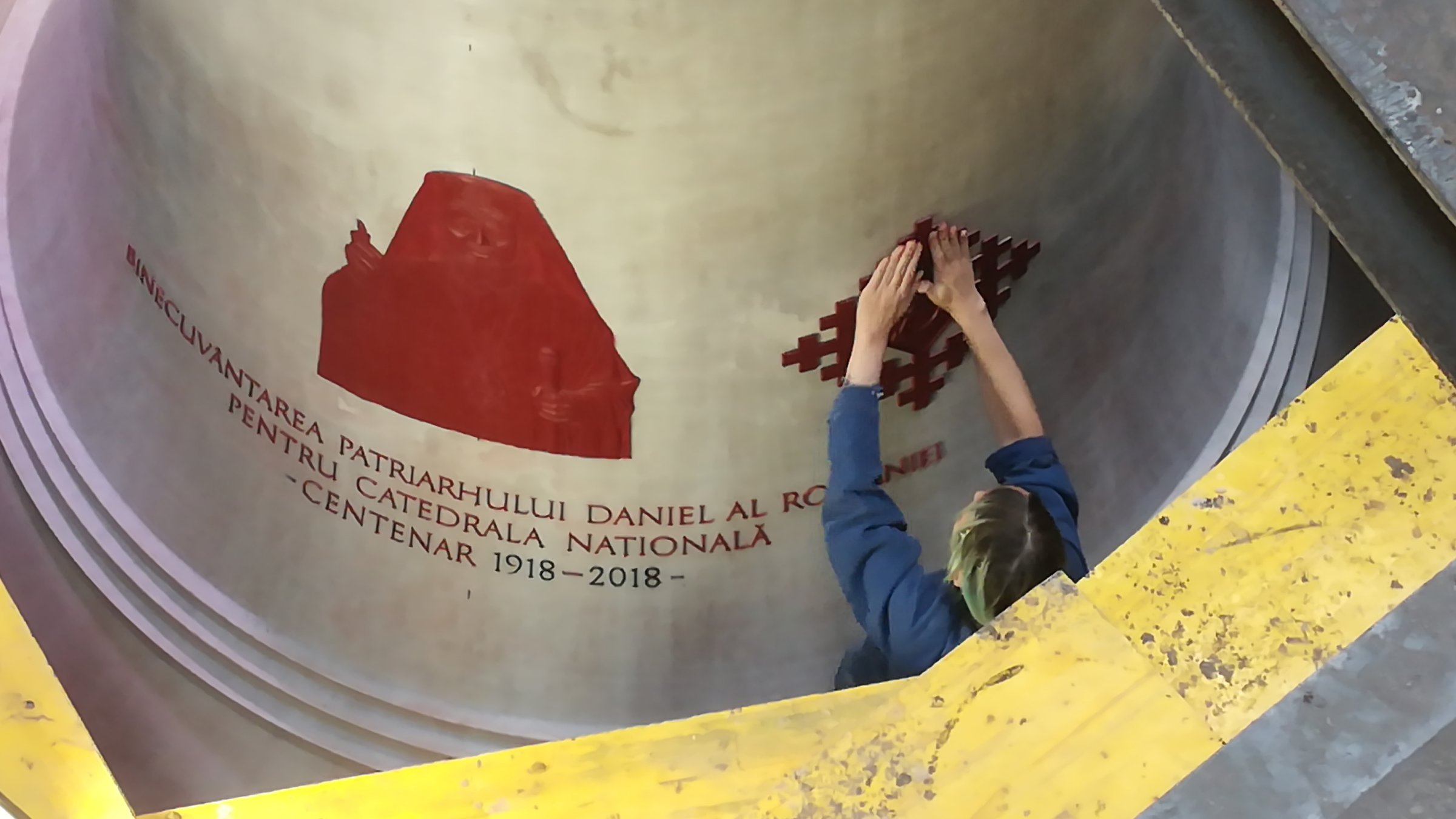 "Eine Glocke für die Ewigkeit": In der Grube der Gießerei steht die riesige wachsbeschichtete „falsche Glocke“ wie mit Zuckerguss übergossen. Vom oberen Rand der Grube sieht man eine Mitarbeiterin, die damit beschäftigt ist, rote Verzierungen an die Glocke zu kleben. An den Außenrändern des Bildes sieht man Teile des Gerüsts, das um die Glocke gebaut wurde.