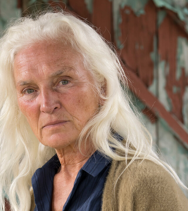 "The Tourist - Irisches Blut - Die Kinder von Lir": Niamh Cassidy (Olwen Fouéré) ist bekleidet mit einem dunkelblauen Hemd und einer offenen, beigefarbenen Fleecejacke. Sie steht vor einer alten Holztür, von der an vielen Stellen bereits der rote Lack abgeblättert ist. Sie blickt ernst in Richtung Kamera.