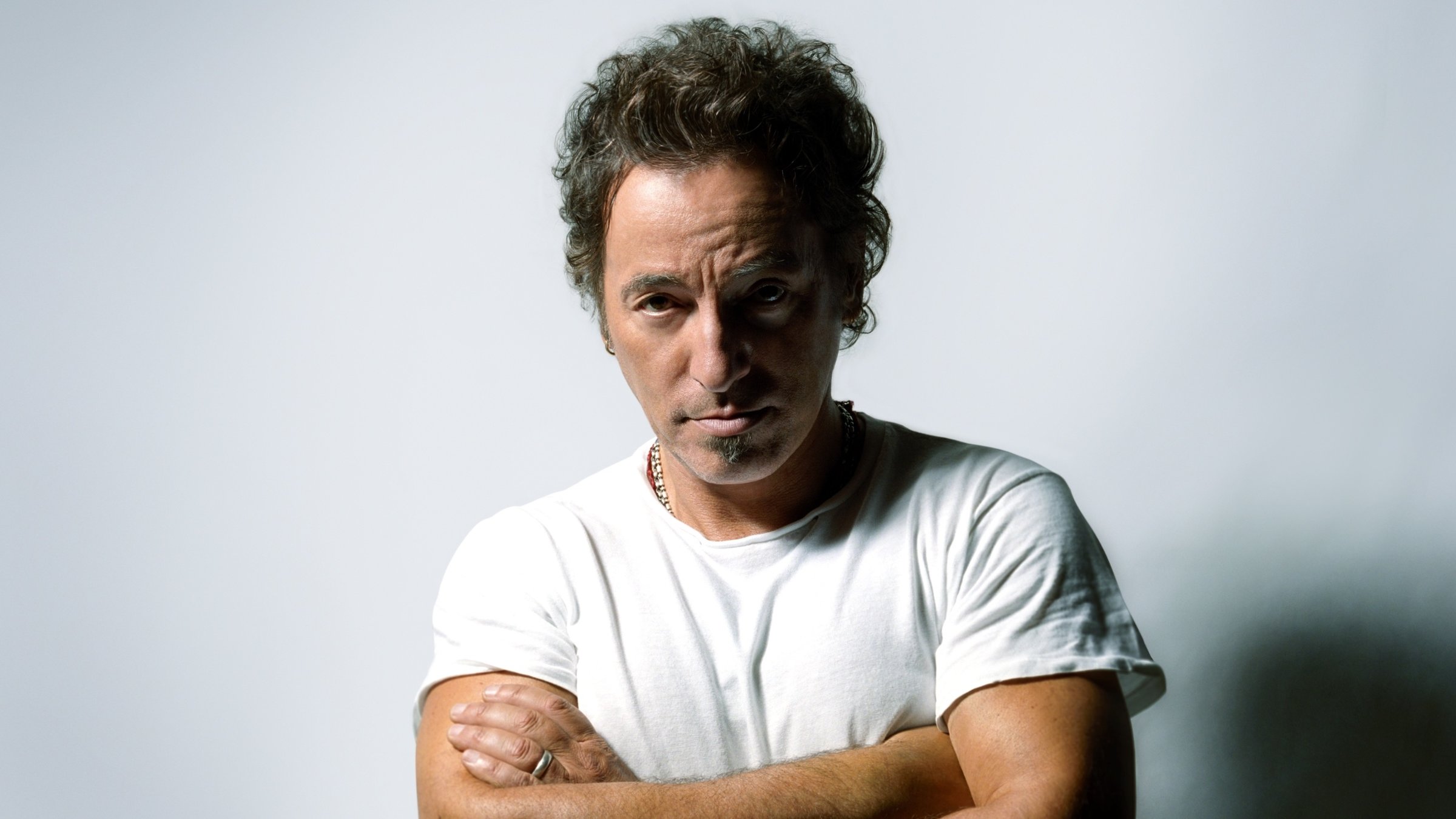 "Bruce Springsteen & The E Street Band: Live in New York City": Bruce Springsteen steht mit verschränkten Armen in einem weißen Tshirt vor einer weißen Wand und schaut direkt in die Kamera.