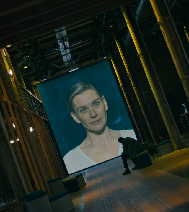 "Concordia - Tödliche Utopie (6) - Kein Weg zurück": Juliane Ericksen (Christiane Paul) ist auf eine Leinwand projiziert. Davor sitzt Noah Ericksen (Steven Sowah) in einem dunklen Raum und sieht zu.