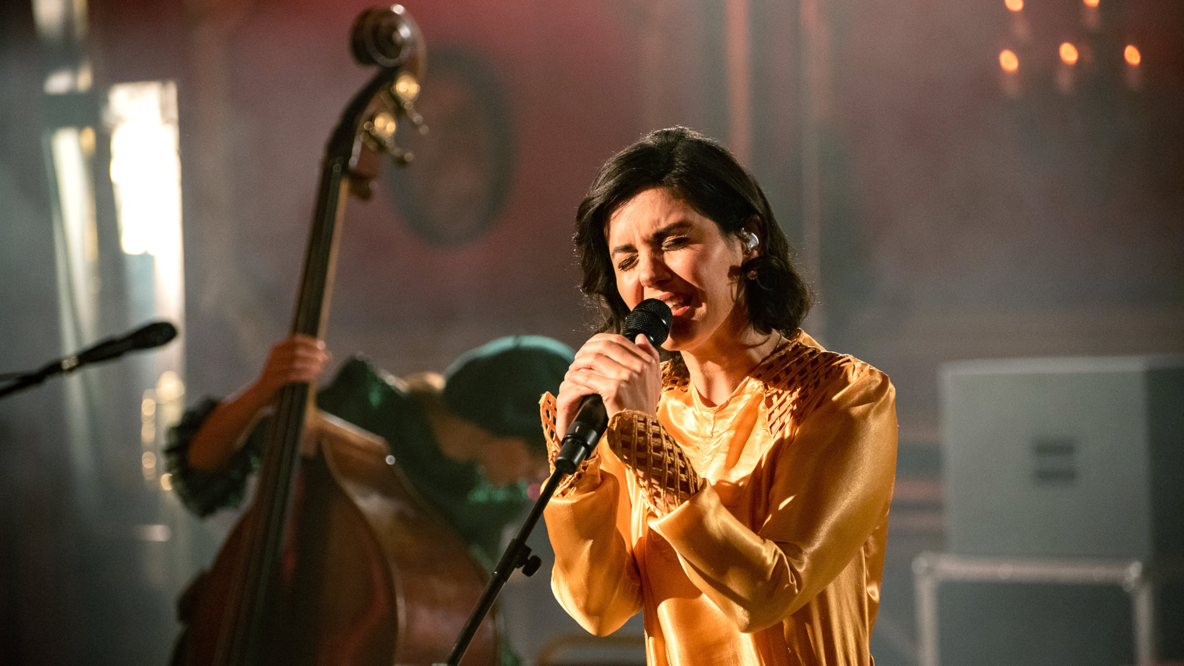 "Katie Melua: From the Rivoli Ballroom": Eine Frau mit schwarzen Haaren und goldenem Kleid hält ein Mikrofon dicht vor den Mund und singt mit geschlossenen Augen. Im Hintergrund ist eine Kontrabass-Spielerin schemenhaft zu sehen, die sich gerader herunterbeugt.