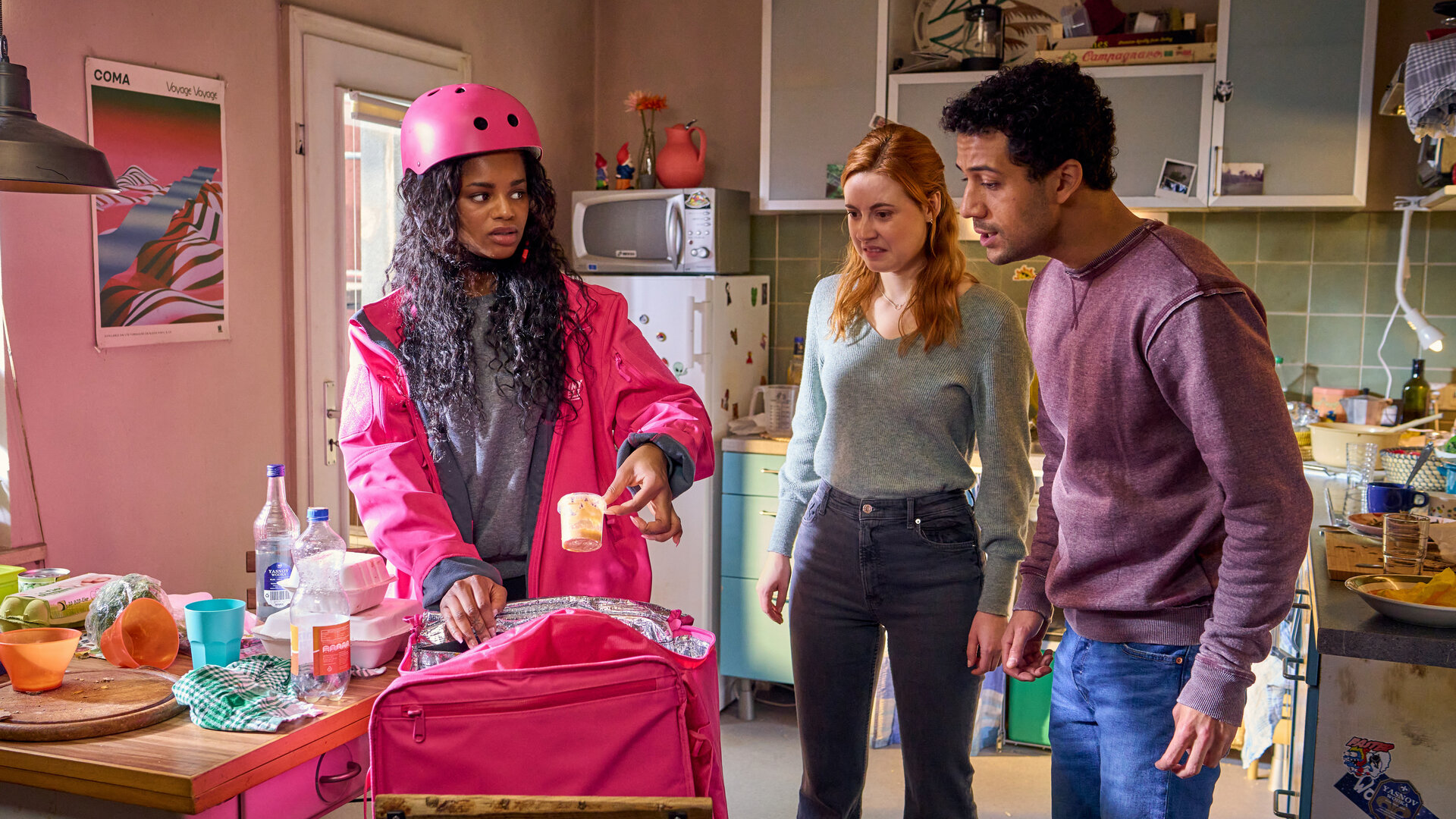 "Ich dich auch! - Der Dreier": Smilla (Melodie Simina), Caro (Meriel Hinsching) Yannik (Rojan Juan Barani) stehen in der Küche. Smilla trägt einen pinken Fahrradhelm. Sie holt Foodboxen aus aus einer pinken Tasche heraus. Caro und Yannik schauen kritisch und angewidert.