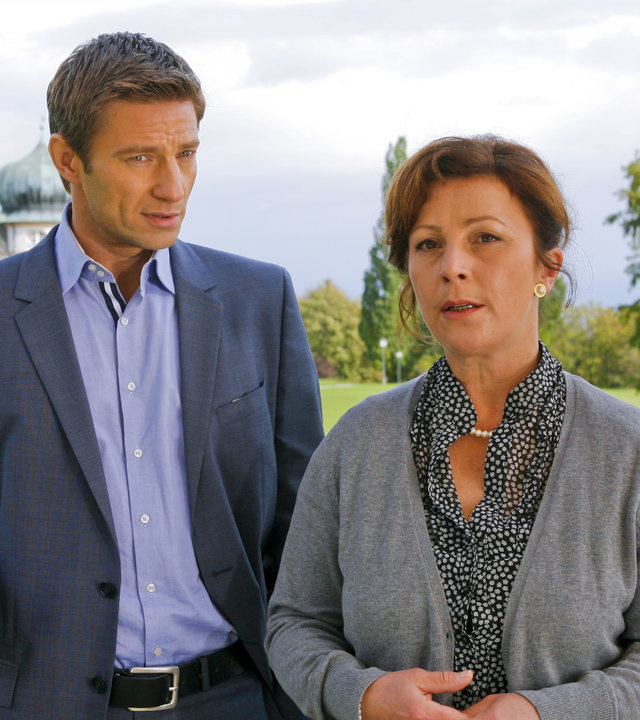 "Die Rosenheim-Cops - Echo des Todes": Auf dem Klinikgelände steht Kommissar Hansen (Igor Jeftic) neben Marianne Eder (Bettina Redlich) und spricht mit ihr. Frau Eder hat den Blick von ihm abgewandt und schaut gedankenverloren nach vorne.