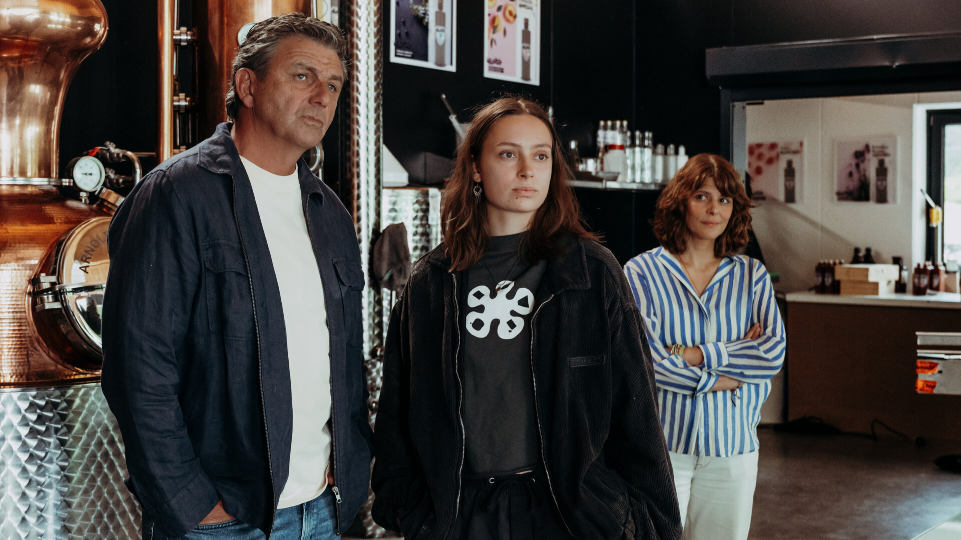 "Der Bergdoktor: Wer einmal lügt": In der Brennerei: Dr. Martin Gruber (Hans Sigl) und Lexi Helmig (Mascha Schrader) stehen nebeneinander vor einem Brennkessel und blicken nachdenklich auf etwas außerhalb des Bildausschnitts. Im Hintergrund steht Isabelle Meyer (Lale Yavaş), die Arme vor der Brust verschränkt, und beobachtet die beiden lächelnd.