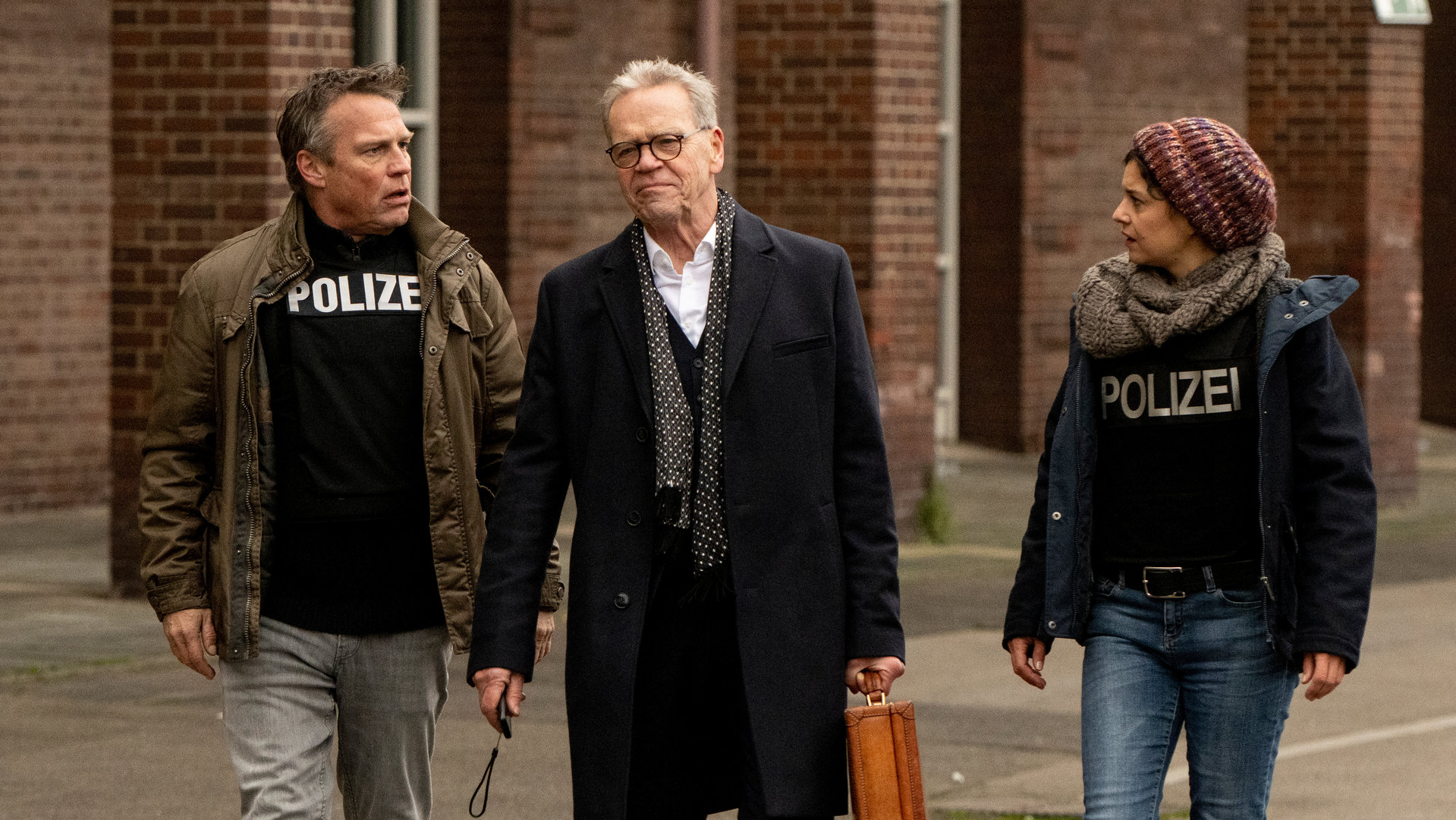 "SOKO Köln - Der Pakt": Auf einer Straße laufen nebeneinander Karl A. Wieland (Jürgen Heinrich), Ermittler Matti Wagner (Pierre Besson) und Lilly Funke (Tatjana Kästel). Die Kommissare sehen Karl A. Wieland fassungslos an. Dieser sieht nach vorne und hält einen Koffer in der linken Hand.