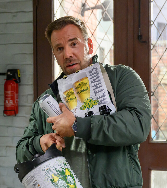 "SOKO Wismar – Kaltgestellt": Lars Pöhlmann (Dominic Boeer) steht in der Wache des Polizeireviers, bepackt mit Bierdosen, Flaschen und einem kleinen Fass der Brauerei Althaus. Durch die Glastür im Hintergrund ist der Hof der Polizeiwache zu sehen.