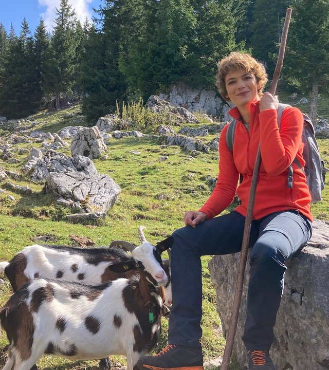 "Terra Xpress - Lockruf der Alpen:" Die Moderatorin Lena Ganschow sitzt auf einem großen Stein auf der Wiese neben Ziegen.