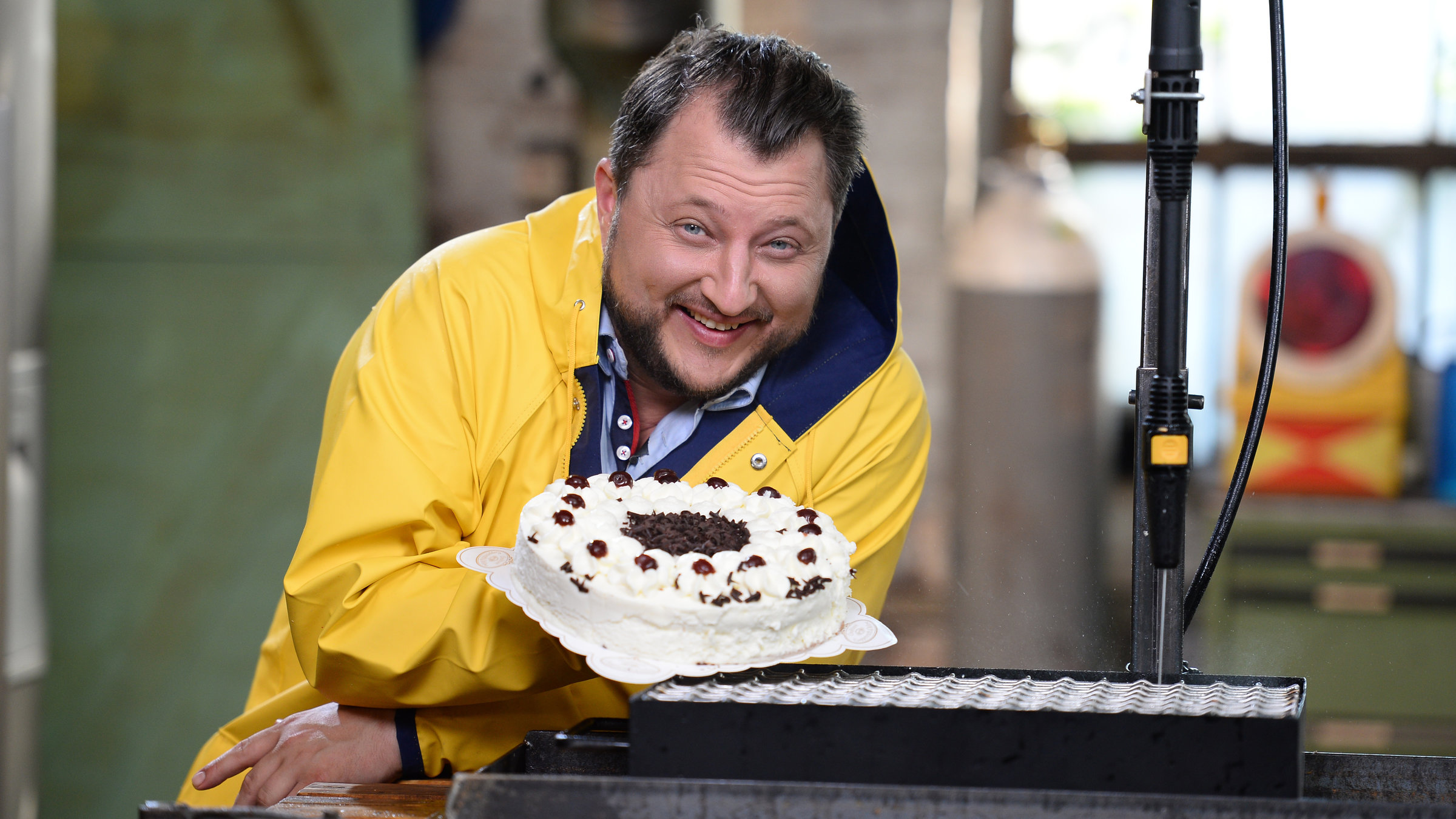 "ZDFzeit: Die Tricks der Lebensmittelindustrie (2)": Sebastian Lege trägt einen Regenmantel und hält eine Sahnetorte in der Hand.