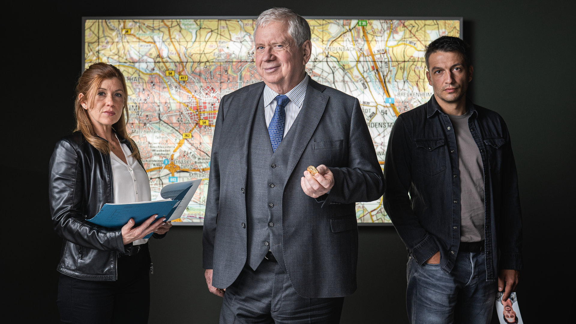 "Der Staatsanwalt - Tödliches Dickicht": Oberstaatsanwalt Bernd Reuther (Rainer Hunold), Hauptkommissarin Kerstin Klar (Fiona Coors) und Oberkommissar Max Fischer (Max Hemmersdorfer) stehen hinter einem Schreibtisch und blicken entschlossen in die Kamera. Reuther hält eine Goldmünze in seiner linken Hand. Auf dem Schreibtisch liegen diverse Unterlagen, im Hintergrund an der Wand ist ein Stadtplan zu sehen.