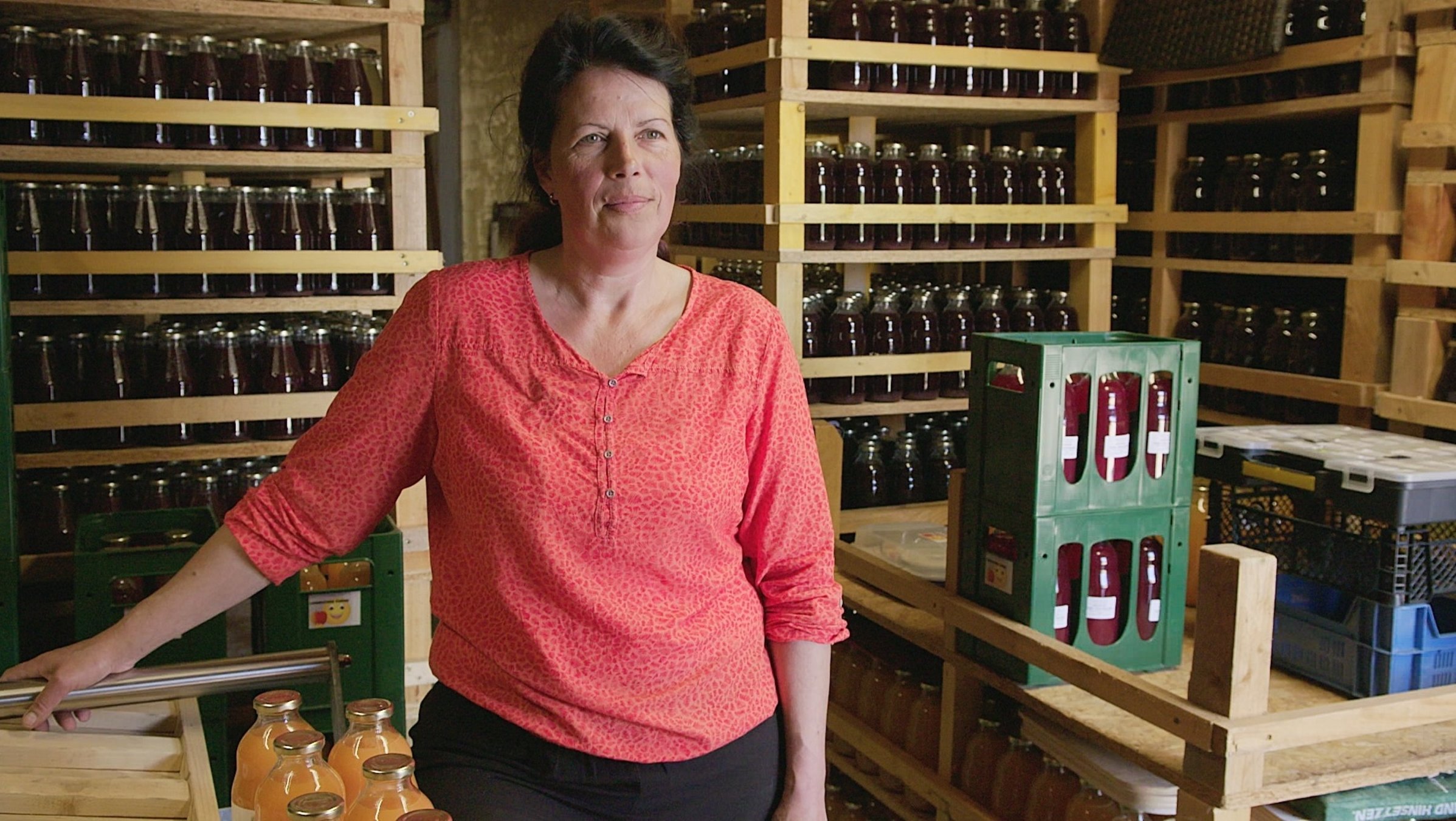 "37°: Hier ist noch lange nicht Schluss - Der Kampf gegen das Dorfsterben": Marlis hat eine orangefarbene Bluse an. Sie steht vor einem Regal mit Apfelsaftflaschen.