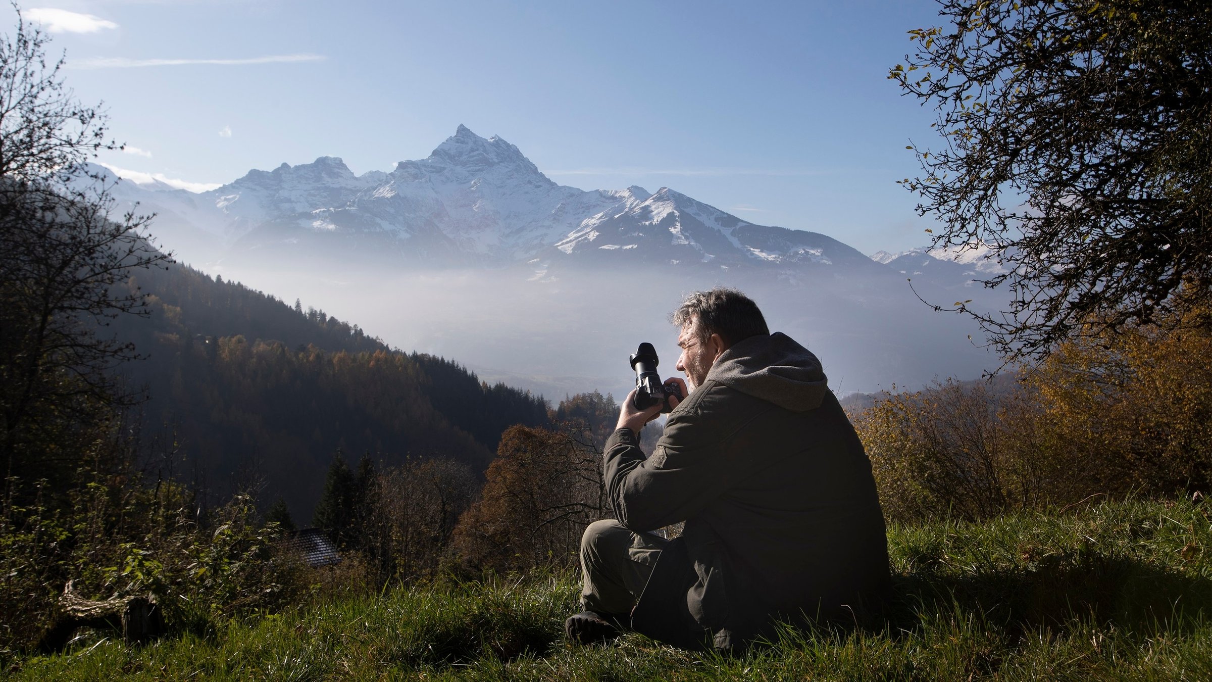 "SCHWEIZWEIT: Leidenschaft Natur – unterwegs in den Waadtländer Voralpen": Pascal Hartwig sitzt im Gras vor einem Alpenpanorama, er hält seinen Fotoapparat in Händen.