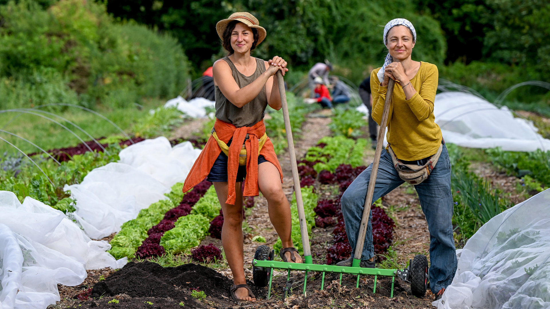 "37° Leben: Anders ackern - Urban Farming": Laura Setzer und Juliane Ranck stehen mit der sogenannten "Campagnole" auf einem Acker zwischen Folientunneln. Verschwommen im Hintergrund arbeiten Freiwillige.