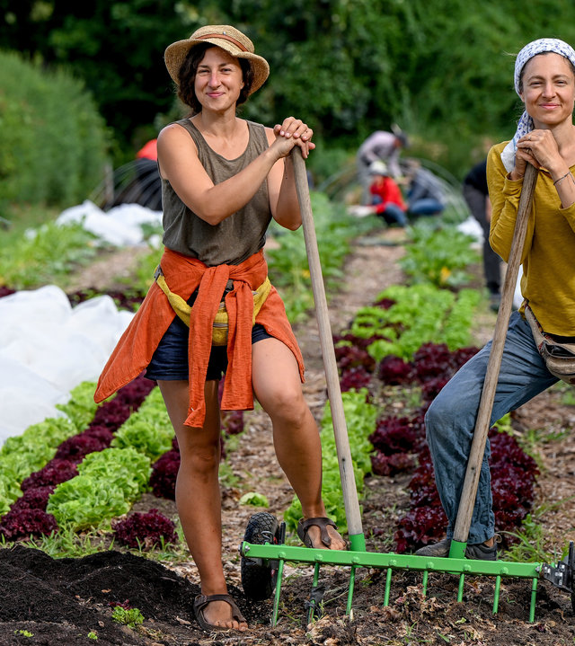 "37° Leben: Anders ackern - Urban Farming": Laura Setzer und Juliane Ranck stehen mit der sogenannten "Campagnole" auf einem Acker zwischen Folientunneln. Verschwommen im Hintergrund arbeiten Freiwillige.