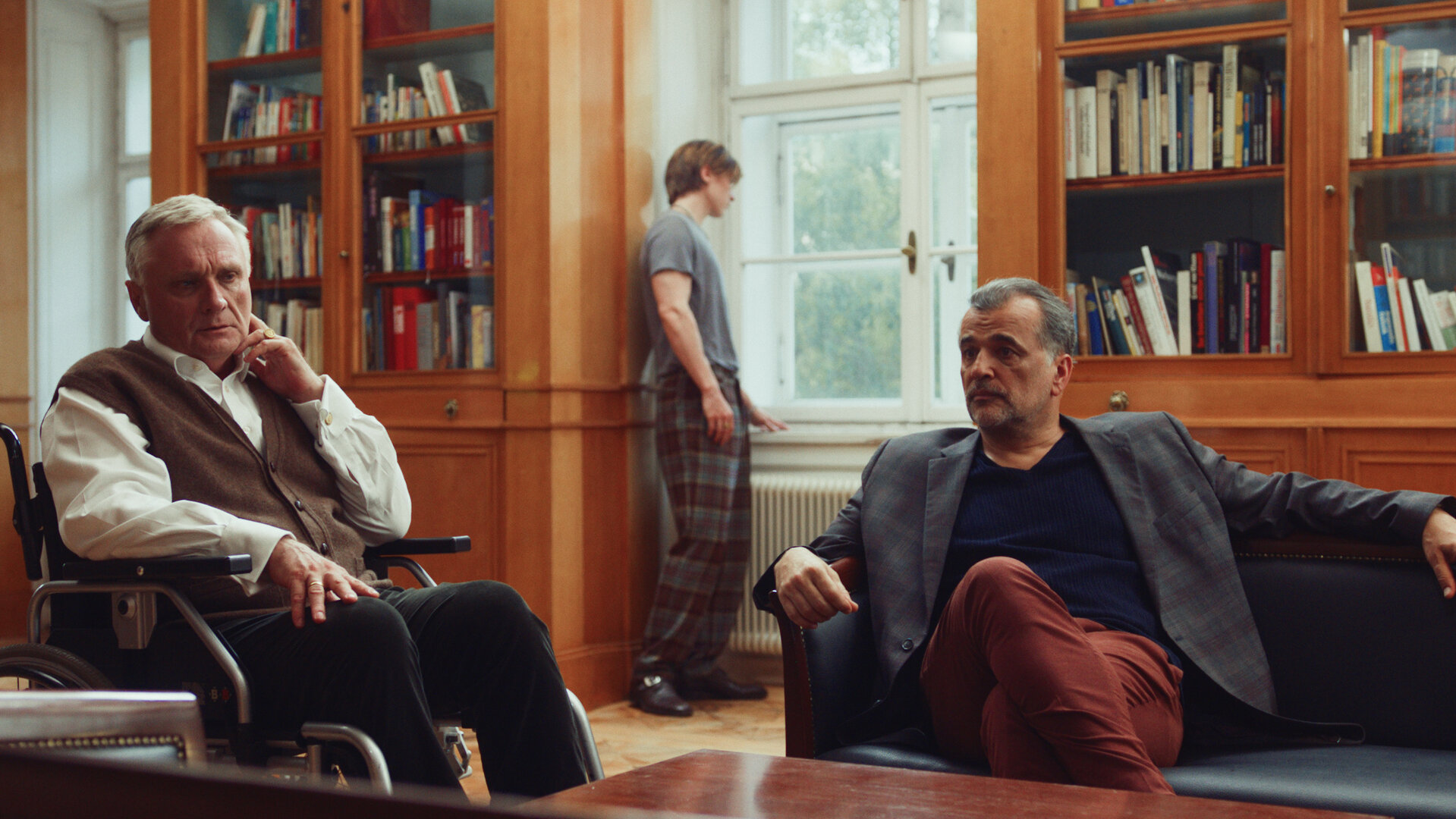 "Crystal Wall - Zwei Seiten": In einem Wohnraum sitzt Bernhard Dardenne (Bernhard Schir) im Rollstuhl, daneben Marc (Michael Masula) auf einem Sofa hinter einem Holztisch. Beide blicken ernst. Im Hintergrund steht Nicolas (Gustav Schmidt) an einem Fenster neben großen Büchervitrinen und schaut nach draußen.