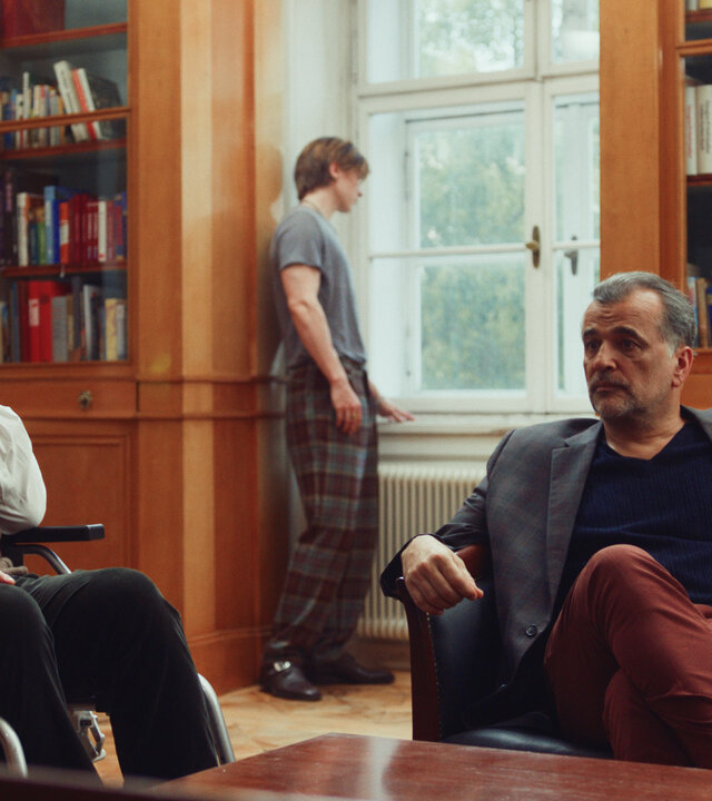 "Crystal Wall - Zwei Seiten": In einem Wohnraum sitzt Bernhard Dardenne (Bernhard Schir) im Rollstuhl, daneben Marc (Michael Masula) auf einem Sofa hinter einem Holztisch. Beide blicken ernst. Im Hintergrund steht Nicolas (Gustav Schmidt) an einem Fenster neben großen Büchervitrinen und schaut nach draußen.